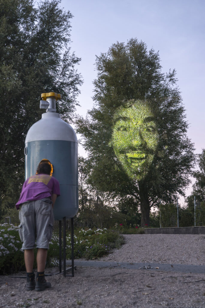 Der niederländische Künstler Jasper Toeli macht mich seinem Kunstwerk „Be the tree“ auf die lebensnotwendige Beziehung zwischen Menschen und Pflanzen. Während Besucher:innen ihren Kopf in einen Sauerstofftank halten, wird ihr Gesicht auf der Krone des gegenüberliegenden Baumes projiziert.
(Foto: Jasper Toeli)