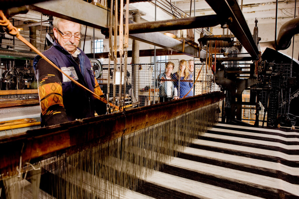 Ein Maschinenvorführer am Webstuhl. Man kann den lautstarken Betrieb der Webstühle hautnah erleben – und viele Fragen stellen.
Bild: LVR-Industriemuseum