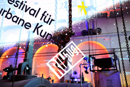 Logo des RUbug Festivals mit buntem Hintergrund in einer alten Halle mit Haken an der Decke Foto: Stefan Juergele