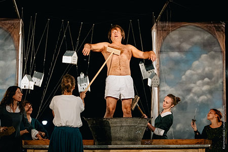 Das Stück Kaspar der polnischen Theatergruppe Teatr Biuro Podrozy: ein Mann wird von umstehenden Frauen mit Besen gewaschen, im Hintergrund hängen an Fäden Miniaturgebäude Foto: Nata Korenovskaya