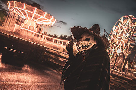 Halloween im Movie Park: ein als Monster verkleideter Haunter steht in Vordergrund, dahinter sind Fahrgeschäfte des Parks wie Riesenrad und Kettenkarussel in düsteren Farben Foto: Movie Park Germany